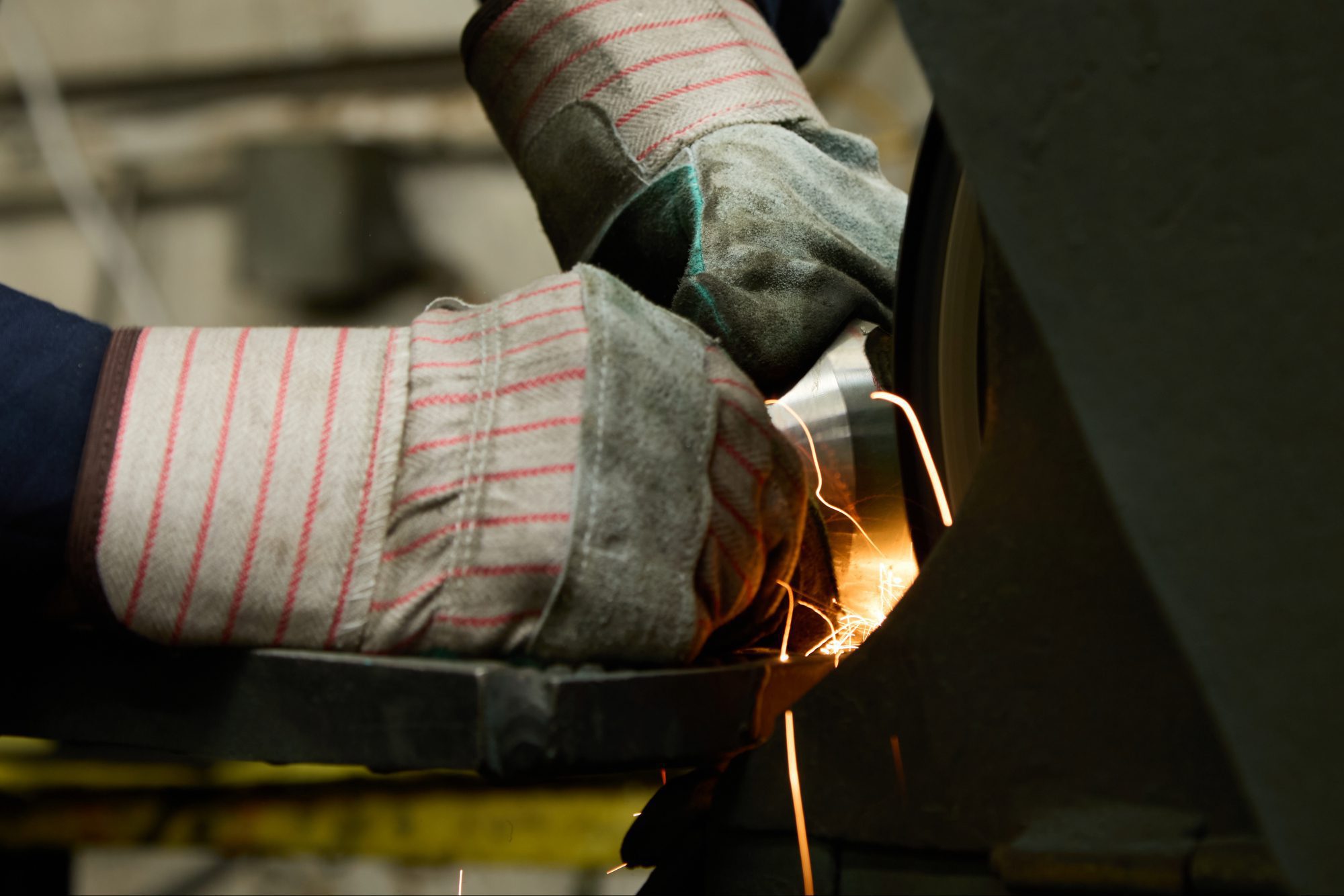 A person wearing thick protective gloves uses a grinder, creating bright orange sparks as they work on a metal object during special product testing in an industrial setting.