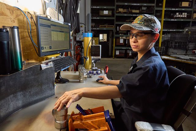 A person wearing safety glasses and a cap sits at a desk in an industrial workspace, using a computer to review material grades. Equipment and metal parts are on the desk, with shelves stacked with boxes in the background.