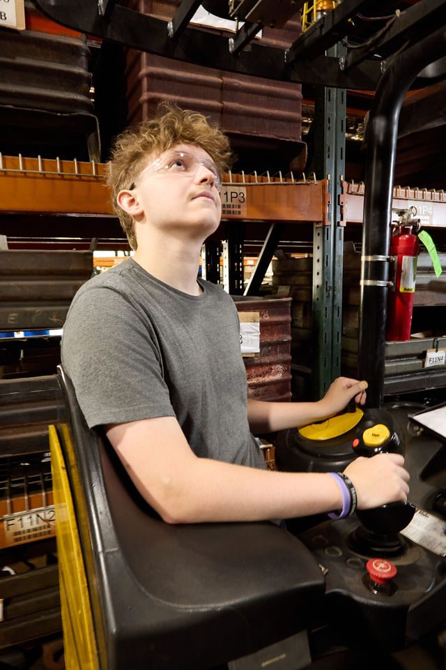 A person wearing safety glasses and a gray t-shirt operates machinery in a warehouse, looking upward. Box quantity packaging is stored on shelves filled with containers and a fire extinguisher is visible in the background.