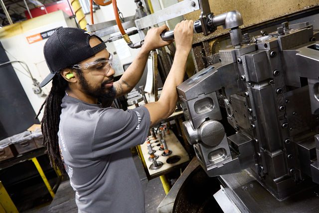 A man wearing safety glasses and earplugs operates heavy machinery in a factory, adjusting custom fittings on a metal component while standing next to an industrial machine with various levers and parts.