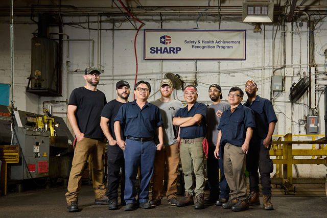 A group of nine workers in uniforms stand together in a factory, smiling at the camera. Above them is a sign reading SHARP Safety & Health Achievement Recognition Program.